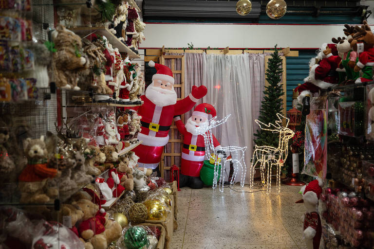 Foto interna de loja na cidade de São Paulo que vende decorações natalinas. Podemos ver uma estante com objetos a venda, como ursos, bengala de açúcar, papais noéis. Ao fundo, dois papais noéis infláveis acenando e uma rena acesa.
