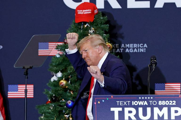 Homem idoso loiro (Trump) de lado, usando terno escuro e gravata vermelha, gesticula, em frente a uma árvore de Natal e cartazes da campanha de Donald Trump 