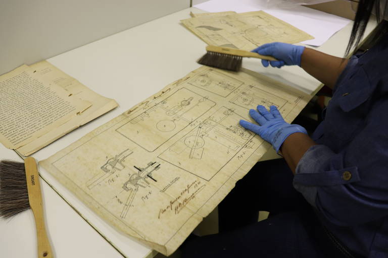 Imagem mostra um documento antigo, uma patente, sendo limpa com uma escova com cerdas macias por uma funcionária negra do INPI, que veste luva azul de borracha