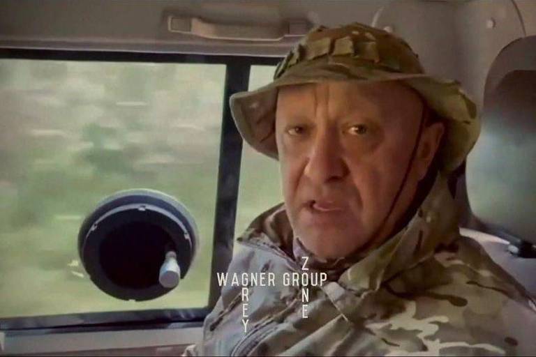 Homem idoso de roupas camufladas e chapéu camuflado fala sentado dentro de um carro