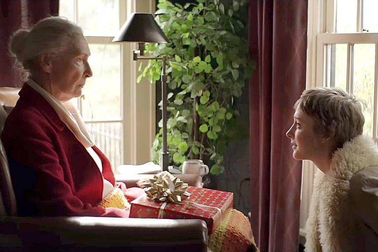 Imagem colorida mostra uma mulher idosa branca sentada em uma poltrona com o olhar distante; em sua frente, uma moça jovem está agachada olhando para ela e sorrindo