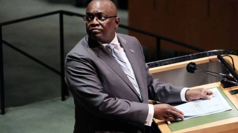 Mokgweetsi é um homem negro e careca; ele usa óculos de grau e veste terno cinza com gravata em um tom de cinza mais claro que o terno e camisa branca; ele está em um púlpito
