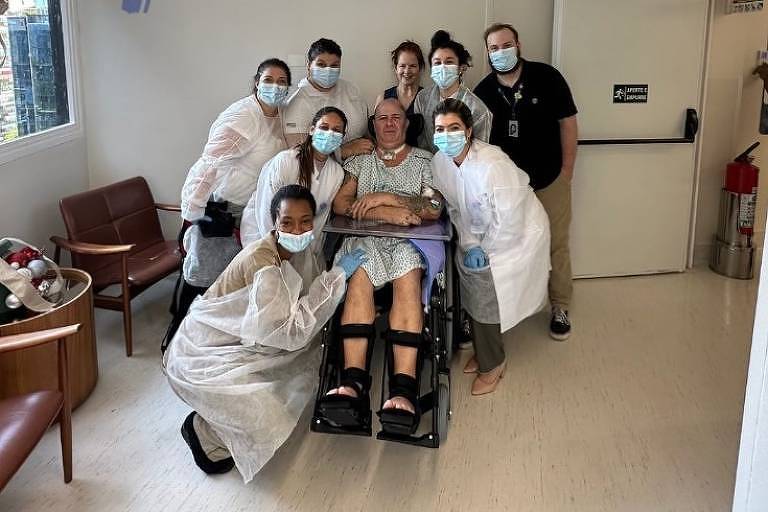 Em foto colorida, homem sentado em uma cadeira de rodas aparece cercado por város médicos em um quarto de hospital