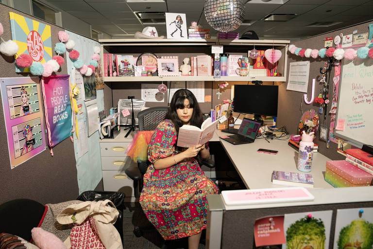 Uma mulher branca de cabelo preto lê um livro em seu cubículo decorado, no escritório onde trabalha