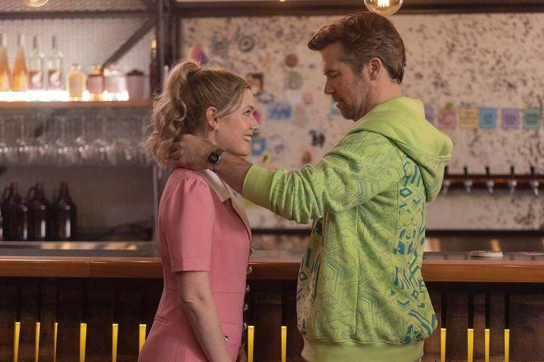 Cena de uma série com um homem e uma mulher, um diante do outro; o homem veste agasalho verde e a mulher veste blusa cor-de-rosa; o homem segura os ombros da mulher