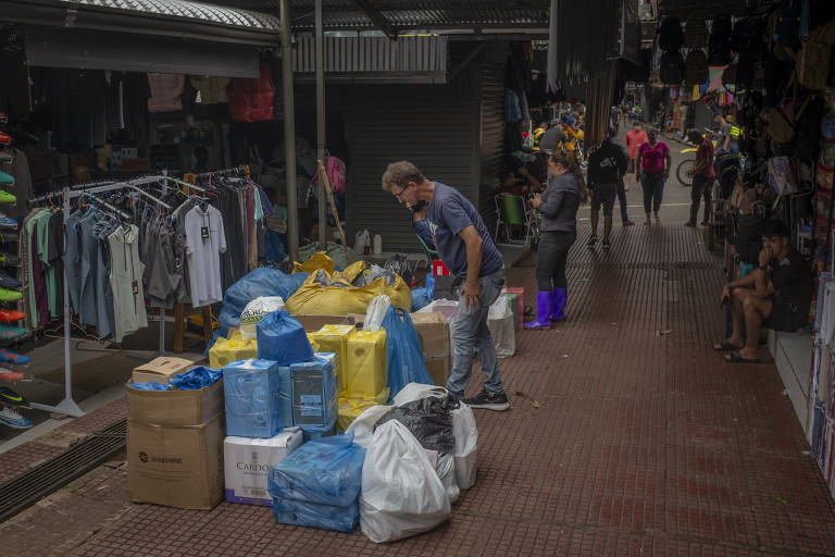 Imagem mostra caixas e sacolas nas cores branca， azul e amarela na rua， com um homem observando