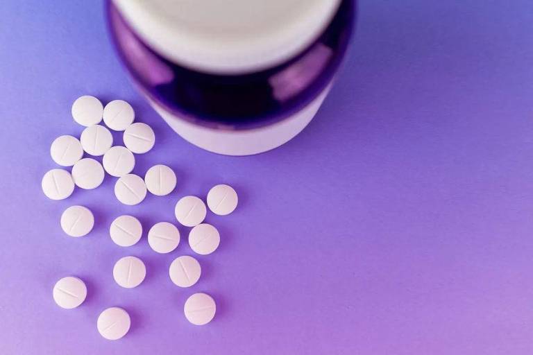 Imagem de um pote de comprimidos, com vários comprimidos para fora sobre uma superfície lilás