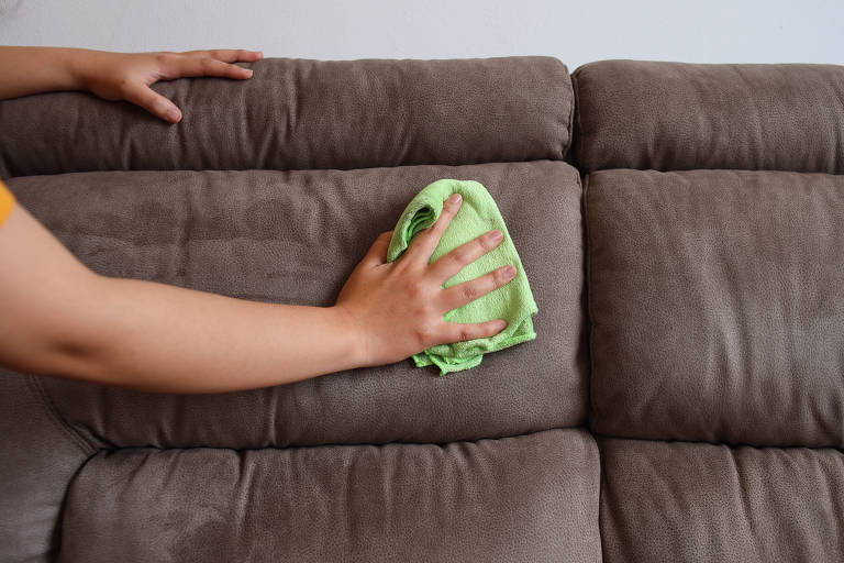 Com um pano verde, uma mão limpa o encosto de um sofá de tecido cinza. A outra mão da pessoa está apoiada na parte superior do sofá