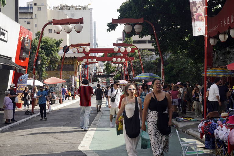 Pessoas usando guarda-chuvas para se proteger do sol caminham por rua do bairro da Liberdade, decorada com lanternas orientais