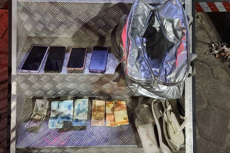 Imagem mostra quatro celulares enfileirados, notas de real e dólar, uma bolsa de acessórios pessoais e um calçado. O material foi apreendido pela polícia militar do Rio durante o Réveillon de Copacabana 