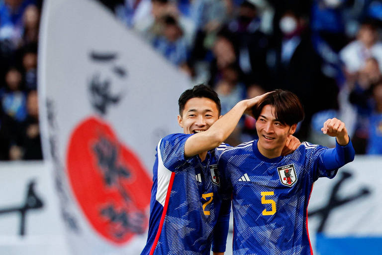 Com a bandeira japonesa ao fundo, Sugawara (camisa 2) e Kawamura (camisa 5) comemoram abraçados gol no amistoso entre Japão e Tailândia em Tóquio