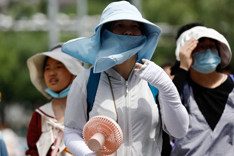 Três mulheres asiáticas usam chapéus de abas largas, lenços e máscaras cobrindo o rosto e blusas e manga comprida para se proteger do sol. Uma delas segura um ventilador portátil