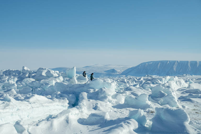 duas pessoas caminham ao longe sobre o gelo, a paisagem de gelo toma conta de toda a imagem, há montanhas ao fundo, céu azul que vai escurecendo até a parte superior da foto