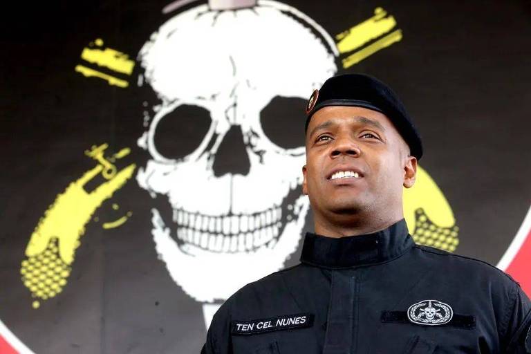 Maurilio Nunes é um homem negro. Ele veste um uniforme do Bope em frente ao símbolo do batalhão de operações especiais: uma caveira com duas pistolas cruzadas atrás