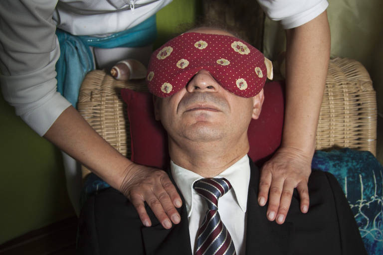 Homem recebe massagem, está vestido com terno e gravata e utiliza um bloqueador de luz nos olhos