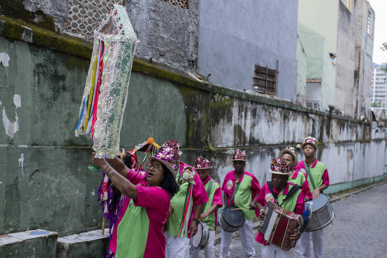 Grupo de pessoas fantasiadas com roupas coloridas, adereços e instrumentos musicais típicos dos folguedos de reisado caminham por viela de comunidade
