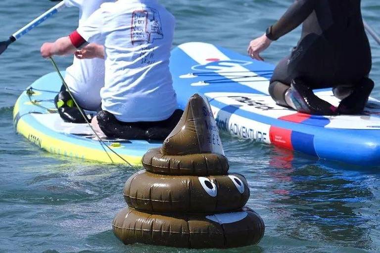 Emoticon de cocô inflável boia na água próximo a pranchas de stand-up paddle
