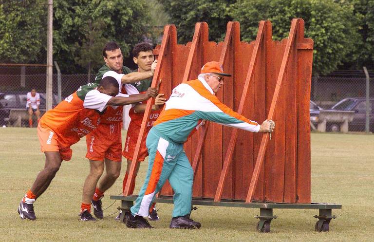 O técnico da Lusa, Zagallo, empurra barreira de madeira auxiliado por jogadores no centro de treinamento do time, no Parque Ecológico do Tietê