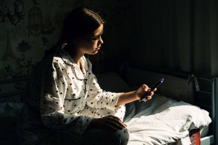 ilustração mostra adolescente segurando celular, ela está sentada na cama e olha para o aparelho
