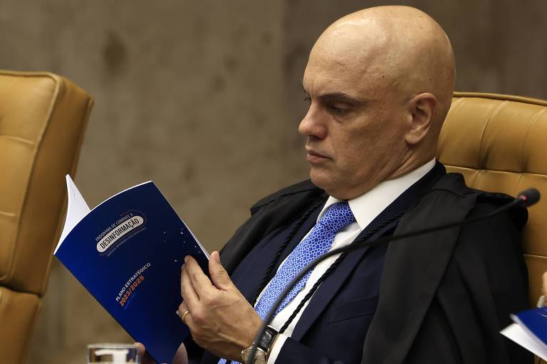 O ministro está sentado em poltrona da corte e folheia livro azul 