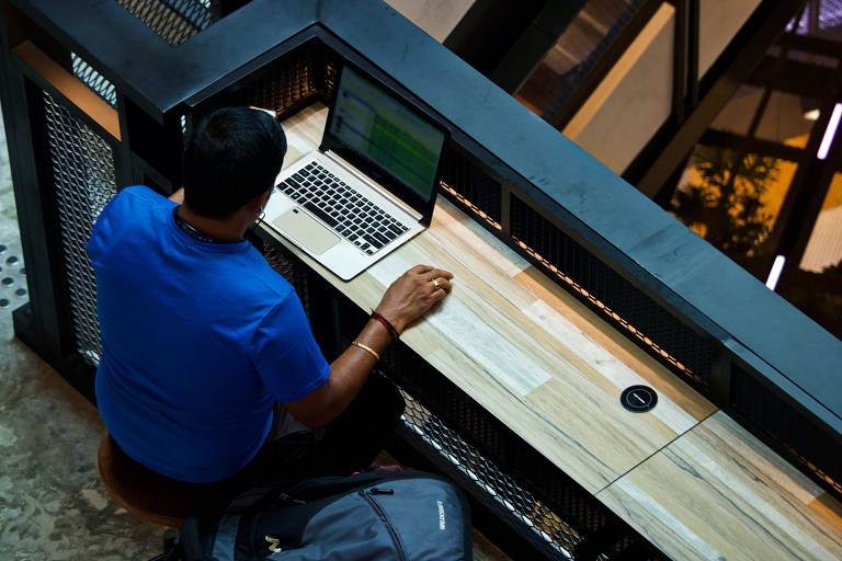 Homem de camiseta azul trabalha em computador sobre uma bancada de madeira