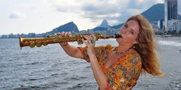 Em foto colorida, A saxofonista, compositora e arranjadora Daniela Spielmann aparece tocando saxofone soprano com uma paraia ao fundo, no Rio de Janeiro