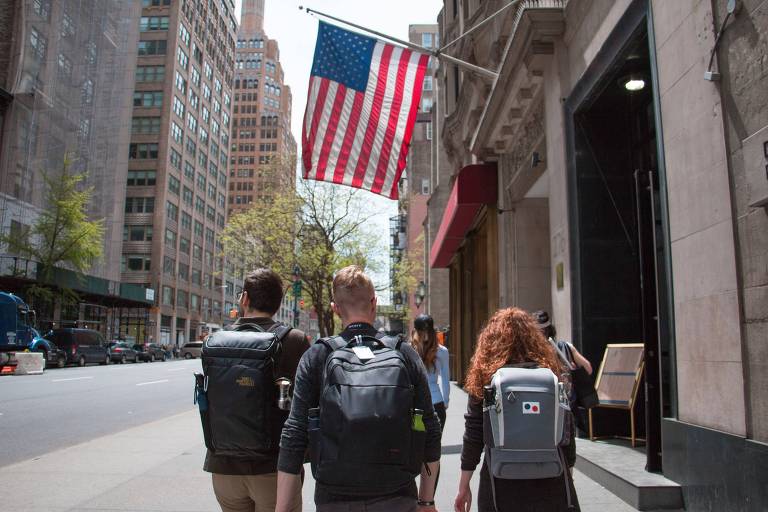 A imagem mostra três pessoas, dois homens e uma mulher, caminhando na rua com mochilas nas costas. Acima delas, há uma bandeira dos Estados Unidos hasteada