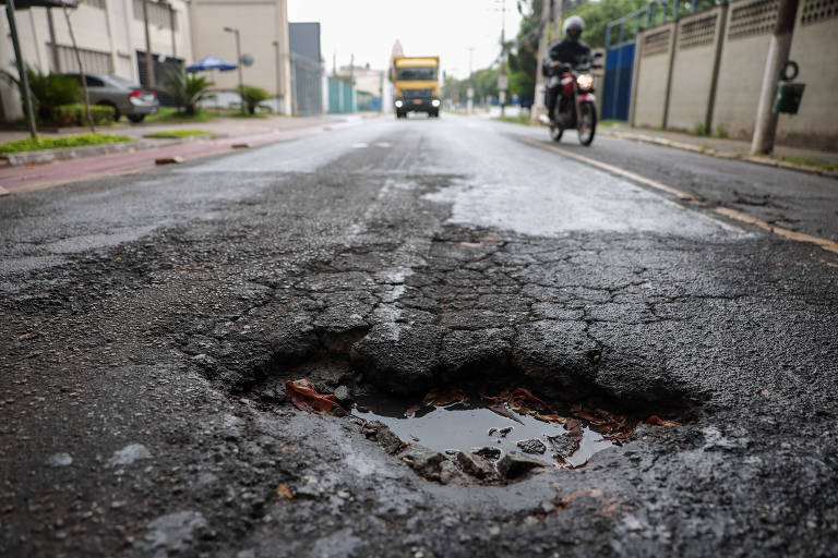Imagem mostra buraco no asfalto, com uma moto e um caminhão ao fundo
