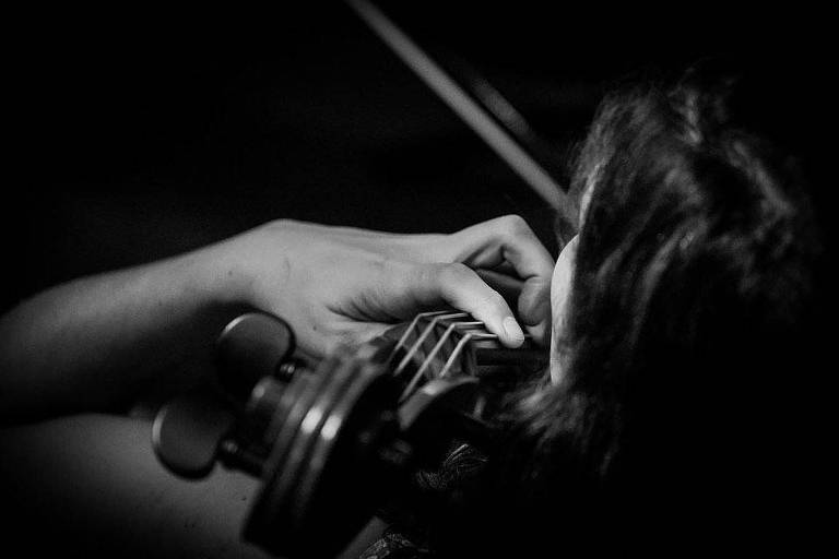 Em foto preto e branco, aparece o detalhe do rosto e da mão esquerda de uma violoncelista tocando o instrumento