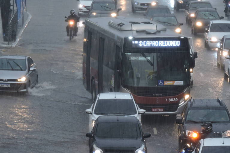 ônibus e carros transitam com faróis acesos por rua alagada 