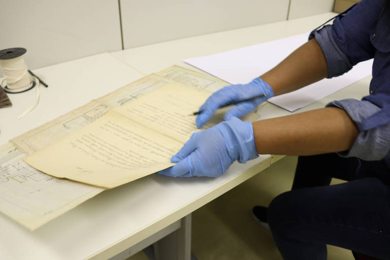 Mãos com luvas azuis seguram documentos antigos, com um lápis em uma delas