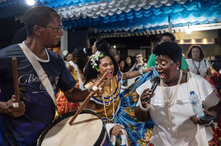 Laura Ramos, mulher negra, canta em festividade. Ao seu redor, mais pessoas dançam.