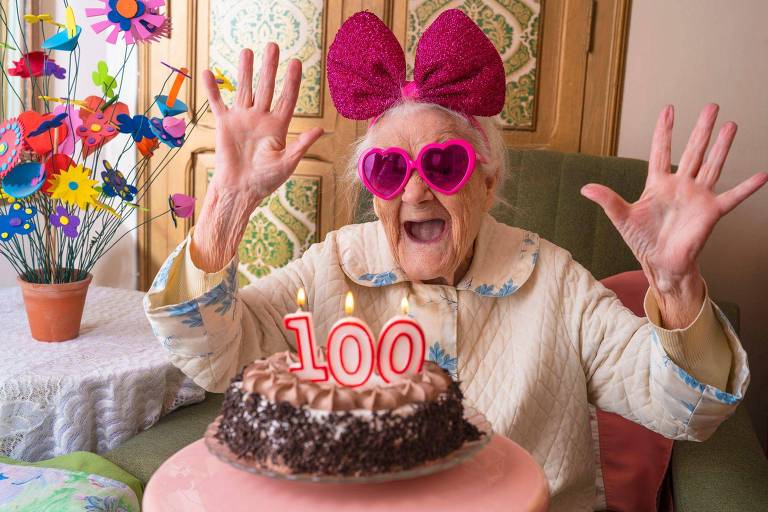 Idosa com óculos de coração e laço rosa em frete a um bolo com velinha de 100 anos

