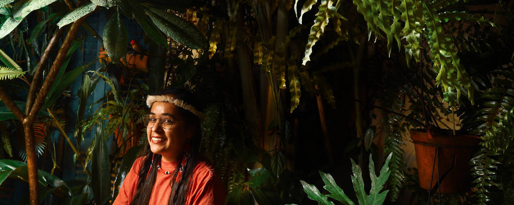 mulher indígena sorri com adereço de penas na cabeça em meio a plantas