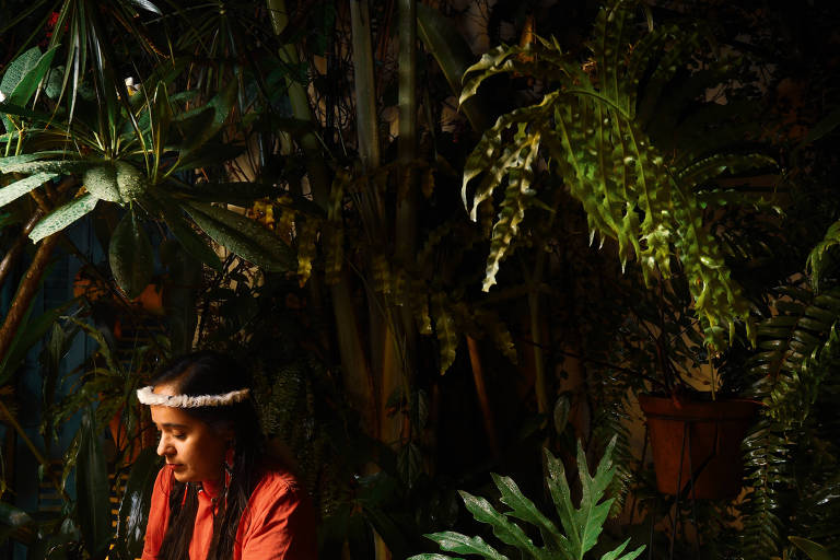 mulher indígena olha para baixo com enfeite na cabeça em frente a plantas