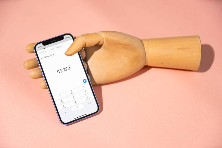 A imagem mostra uma mão de madeira segurando um smartphone com a tela acesa. O fundo é rosa. Na tela do smartphone, há um aplicativo de calculadora aberto 