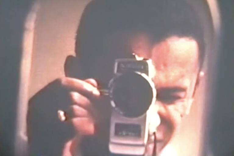 a imagem mostra um homem negro retinto apontando a lente de uma super-8 para um espelho, de modo que a câmera cobre parte de seu rosto