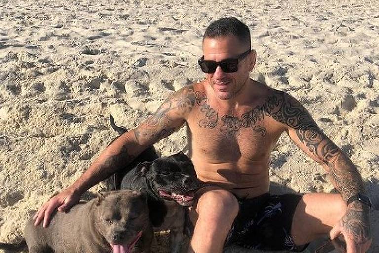 homem sentado na areia da praia sorrindo com um cachorro, sem blusa, bermuda preta e óculos de sol