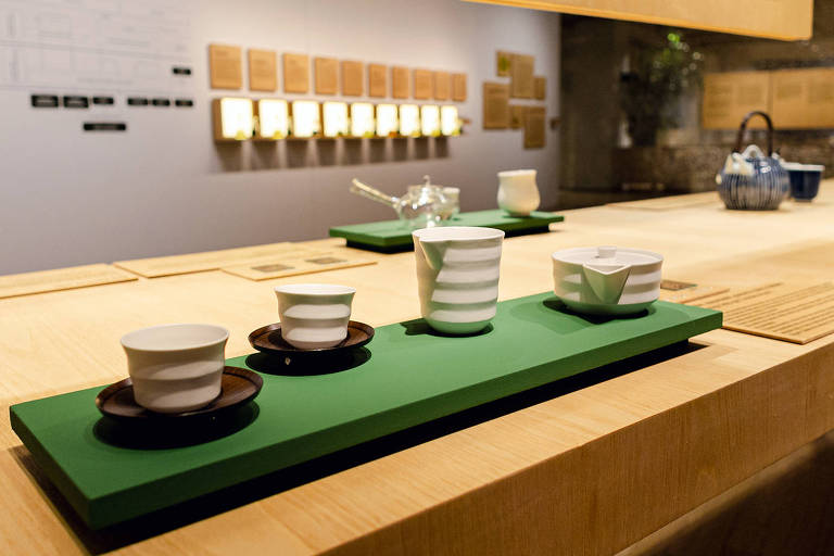 A foto mostra utensílios utilizados no serviço do chá como xícaras e bules sobre uma base verde