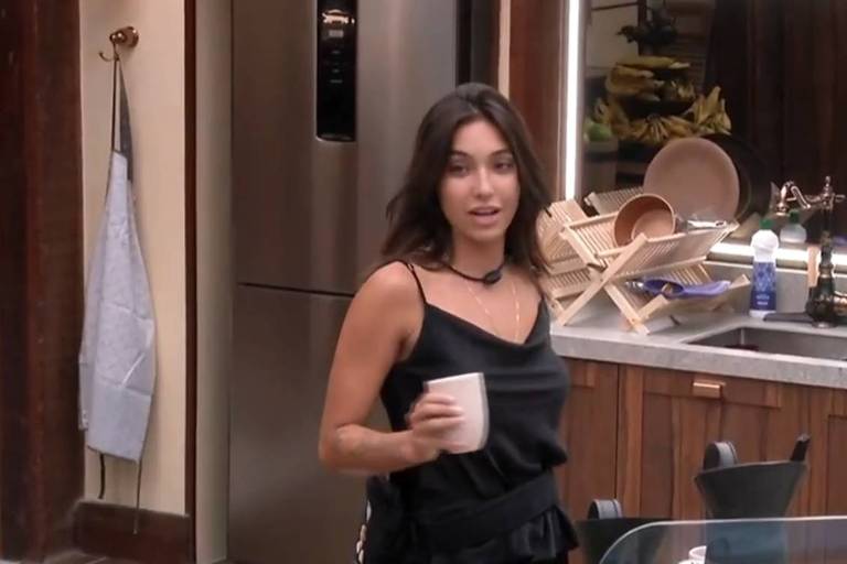 Em foto colorida, mulher de pijama preto toma café da manhã na cozinha
