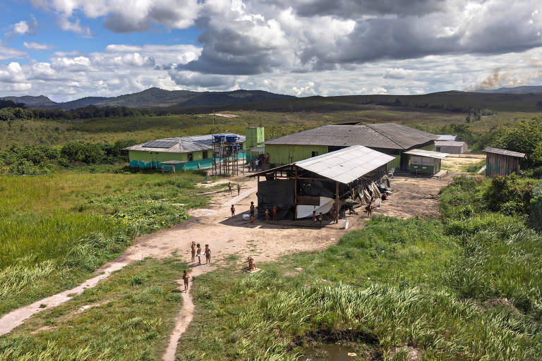 A imagem mostra uma vista aérea de uma comunidade rural com várias construções. No centro, há um grande galpão com um telhado de duas águas e paredes abertas. Ao redor, há casas de cores variadas, incluindo verde e azul. Crianças estão brincando e caminhando em um caminho de terra que atravessa um campo verde. O céu está parcialmente nublado, com nuvens grandes e escuras, e ao fundo, montanhas podem ser vistas.