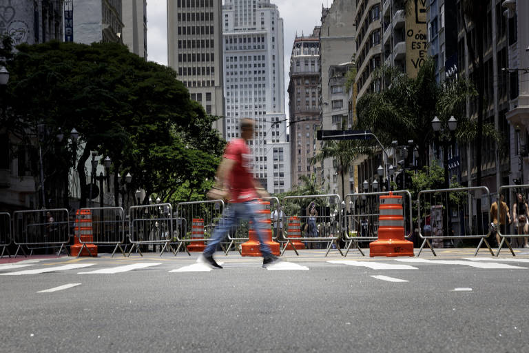 Homem com calça jeans e camiseta vermelha anda furtivamente seobre uma faixa de pedestres no asfalto, ao lado de grandes cones de trânsito laranja e grades de metal; ao fundo se vê o edifício Altino Arantes