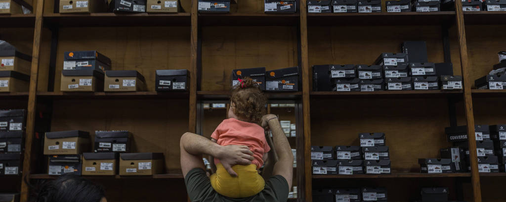 Pai carrega criança nos ombros diante de uma grande estante com caixas de sapatos