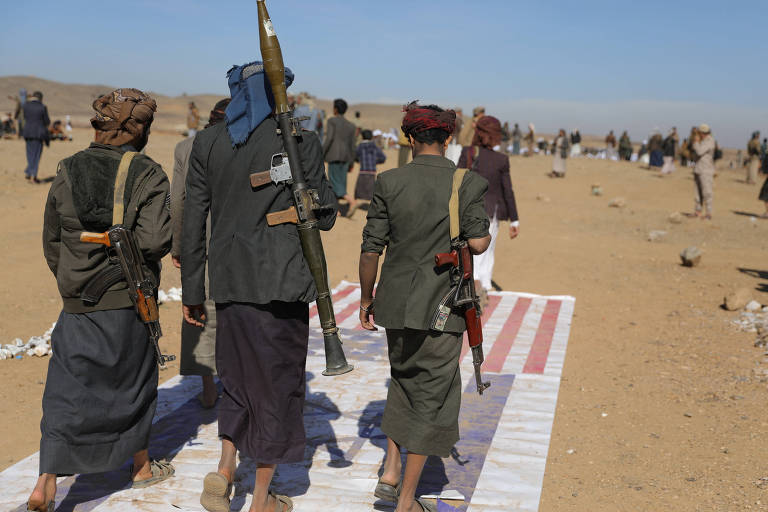 Homens do grupo Houthis caminham sobre as bandeiras de EÛA e Israel em meio a um terreno similar ao de um deserto. Eles carregam fuzis e um lança mísseis nas costas, e trajam saias e turbantes 