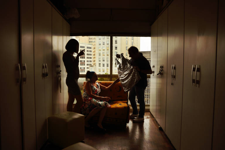 Três pessoas posam para foto em quarto com armários fechados na parede; uma delas está de pé, usando vestido e peruca; outra está sentada em poltrona; a terceira está de pé usando roupas masculinas e segurando um vestido brilhante