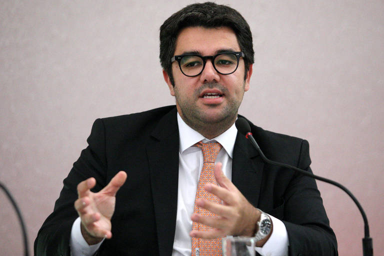 A imagem mostra um homem de óculos, vestindo um terno preto, camisa branca e gravata laranja, falando em uma conferência. Ele está gesticulando com as mãos e há um microfone à sua frente.