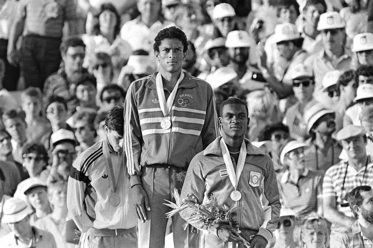 Foto em preto e branco com Joaquim Cruz no topo do pódio, observado por torcedores; o corredor está com a medalha no peito e veste um agasalho