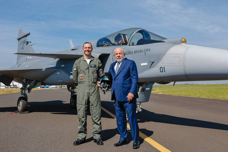Foto mostra o presidente Lula, de terno azul, e um militar da Aeronáutica em roupa de voo na frente de um avião de caça cinza, numa pista de pouso
