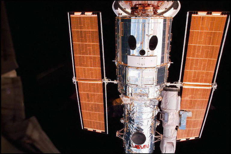 Telescópio Espacial Hubble, um grande cilindro metálico em tom prateado ladeado por duas estruturas cobertas por placas solares