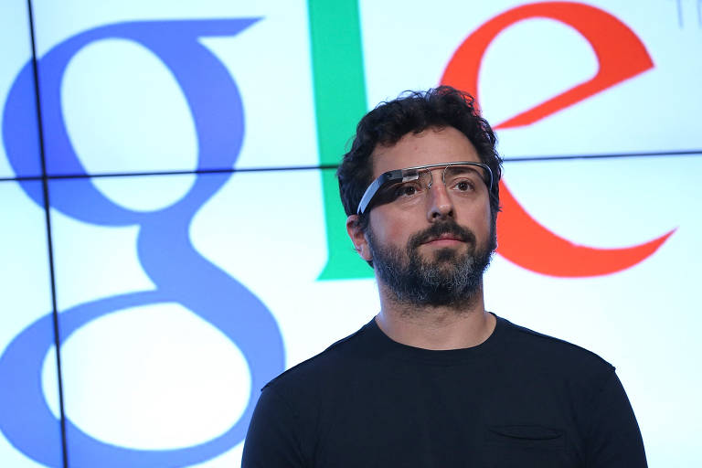 Um homem está usando um dispositivo Google Glass e está em frente a um fundo com o logotipo do Google. O homem tem cabelo escuro e barba， e está vestindo uma camiseta preta. O logotipo do Google é exibido em cores vibrantes， com as letras 039;g039;， 039;o039;， 039;o039;， 039;g039;， 039;l039; e 039;e039; em diferentes cores.
